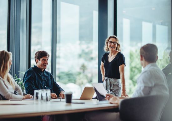 Four coworkers at a boardroom table. One of them is a woman standing casually speaking to the group.